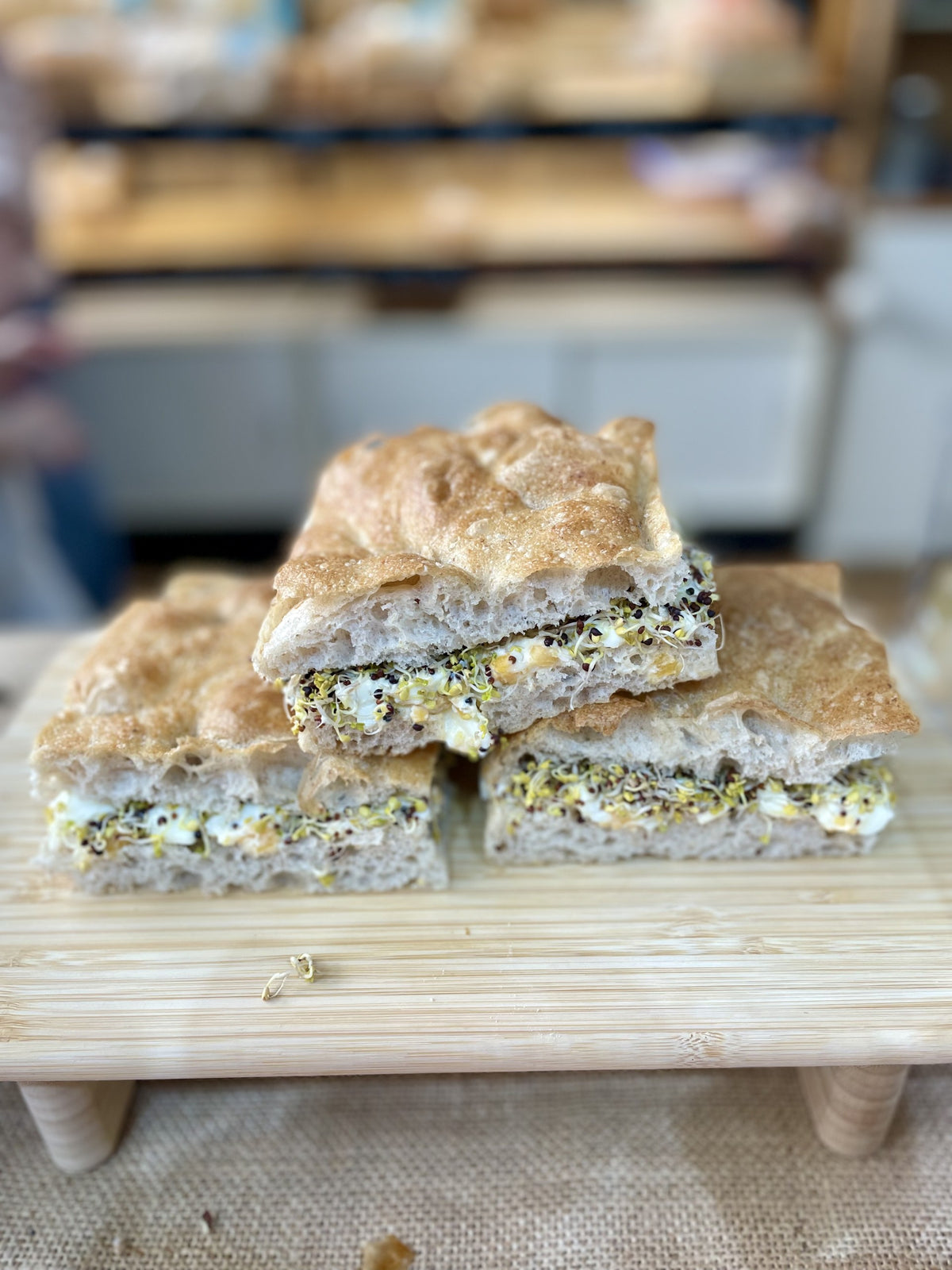 Freshly prepared egg mayonnaise and sprout focaccia sandwiches stacked on a wooden serving board at Wild Oats in Bristol, with a softly blurred shop background.