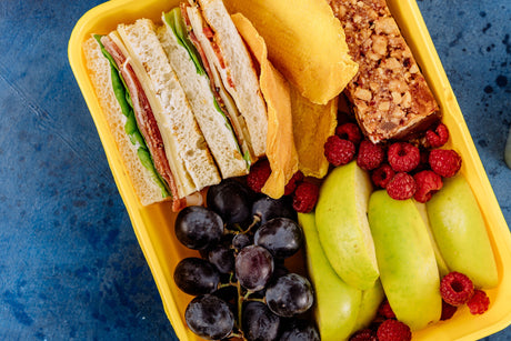 Yellow school lunchbox with sandwiches, apple slices, grapes, raspberries, dried mango and a snack bar
