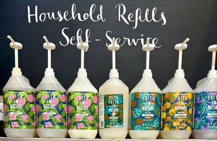 Row of white bottles with colourful 'Faith in Nature' branded labels on a black background with text 'Household Refills Self-Service'.